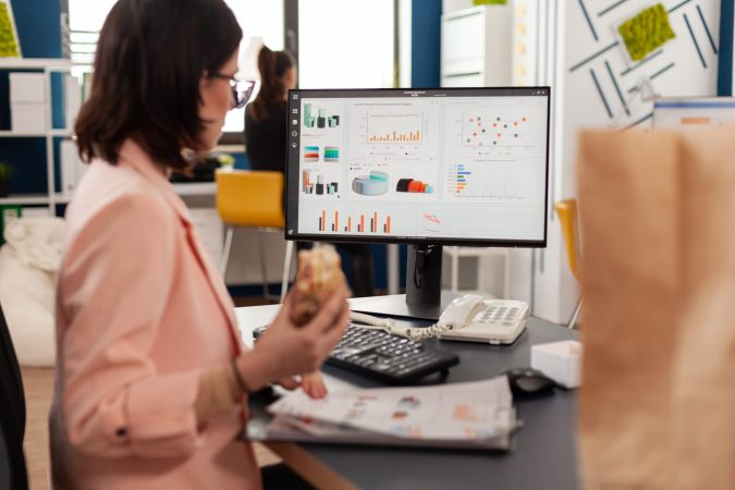 Woman in a pink blazer sitting at a desk, holding a sandwich and looking at a computer monitor displaying various bar charts, pie charts, and graphs.