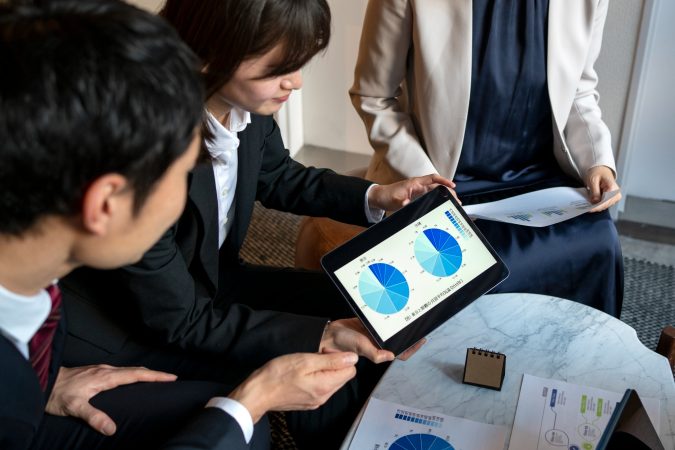 Three professionals in suits sitting around a table, one holding a tablet showing blue pie charts while others look on and discuss.