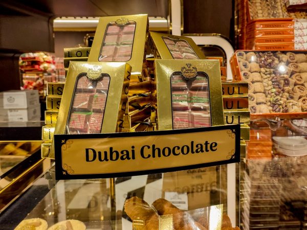 Display of luxury Dubai chocolate gift boxes in gold packaging on a shop shelf, with assorted sweets in the background.