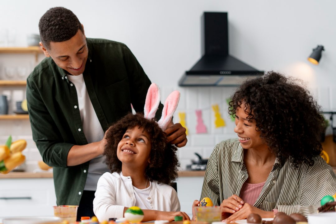 In a bright kitchen, two adults smile as one places pink bunny ears on a child’s head while they sit at a table with decorated Easter eggs and craft supplies.
