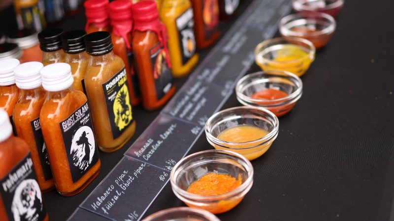A market stall displaying rows of hot sauce bottles and tasting bowls on a black table, with chalkboard labels describing each variety